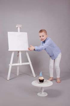 preschool boy in white pants and blue shirt on grey background near easel with mockup and cupcake with candle, shaped in number four 4. happy birthday. celebration. High quality photo