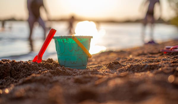 Children's toys for playing on the sand. Plastic bucket and rake on the beach at sunset. The concept of summer, family holidays and vacations.