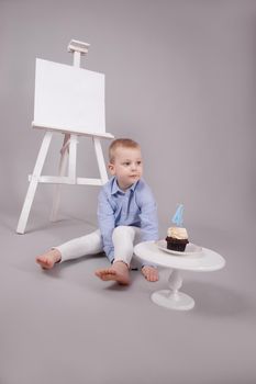 preschool boy in white pants and blue shirt on grey background near easel with mockup and cupcake with candle, shaped in number four 4. happy birthday. celebration. High quality photo