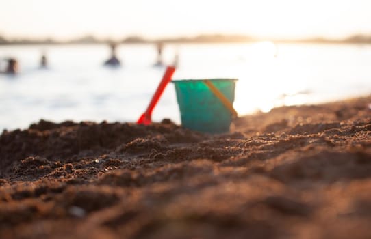 Children's toys for playing on the sand. Plastic bucket and rake on the beach at sunset. The concept of summer, family holidays and vacations. The bucket is out of focus. Focus on the sand