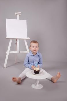 preschool boy in white pants and blue shirt on grey background near easel with mockup and cupcake with candle, shaped in number four 4. happy birthday. celebration. High quality photo