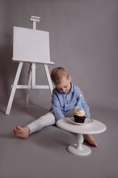 preschool boy in white pants and blue shirt on grey background near easel with mockup and cupcake with candle, shaped in number four 4. happy birthday. celebration. High quality photo