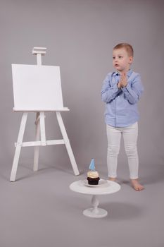 preschool boy in white pants and blue shirt on grey background near easel with mockup and cupcake with candle, shaped in number four 4. happy birthday. celebration. High quality photo