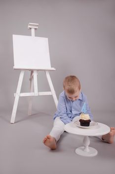 preschool boy in white pants and blue shirt on grey background near easel with mockup and cupcake with candle, shaped in number four 4. happy birthday. celebration. High quality photo