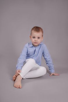 preschool blonde boy in white pants and blue shirt on grey background. child, kid isolated.