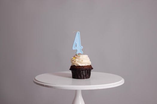 tasty chocolate cupcake with white topping cream and candle in shape of number four 4. on a white stand on a gray background. sweets, bakery. party food. dessert.