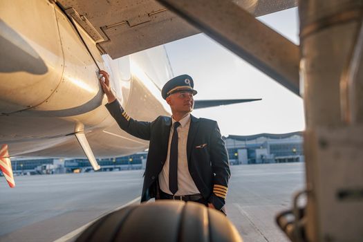 Portrait of professional young male pilot in uniform and hat looking aside, leaning on a big passenger airplane, posing in airport at sunset. Aircraft, occupation, transportation concept