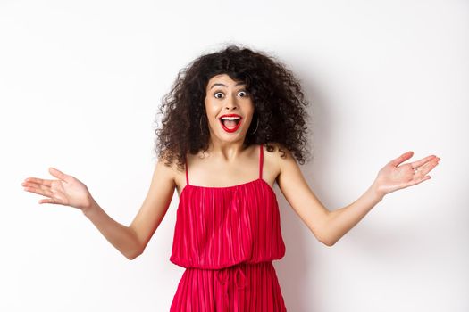 Excited young woman in red dress, spread hands sideways and screaming surprised, standing happy on white background.