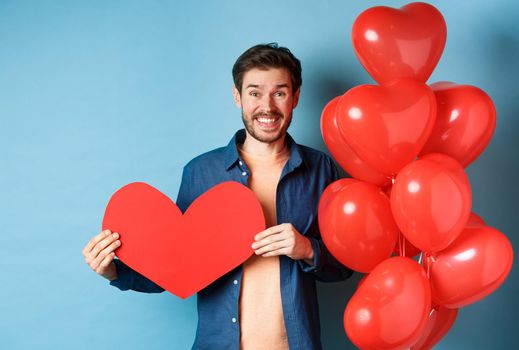 Valentines day concept. Smiling man say I love you, holding paper red heart cutout, standing near romantic balloons, blue background.