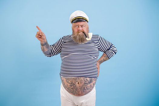 Plus size man with tattoos on large tummy in sailor suit with ancient smoking pipe points aside by finger on light blue background in studio