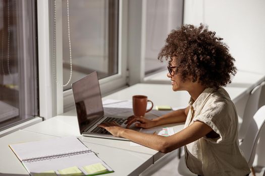 Smiling concentrated female administrative typing messages on laptop while sitting near the window in the office