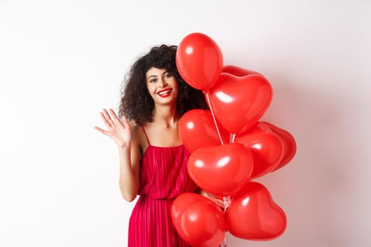 Beautiful lady with curly hair, standing near valentines holiday balloons and waving hand, saying hello, standing against white background.