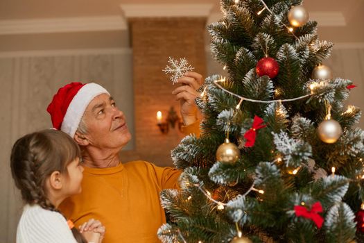 Mature man in santa claus hat and orange shirt looking at decorating fir tree and holding granddaughter on hands, kid and grandpa posing in living room.