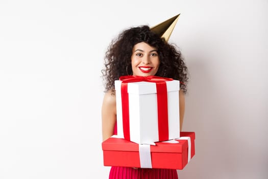 Holidays and celebration. Beautiful lady with curly hair, wearing party hat and holding big presents, smiling happy, receiving birthday gifts, standing on white background.