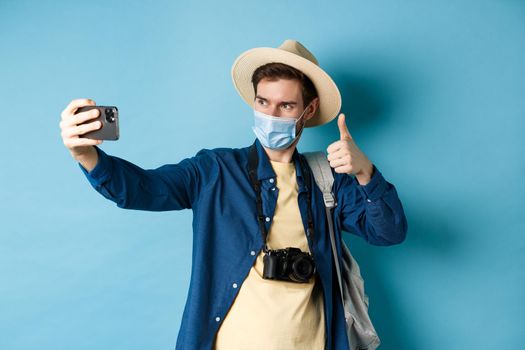 Covid-19, pandemic and travel concept. Happy male tourist taking pictures on vacation, make selfie with thumb up, recommending hotel, standing on blue background.