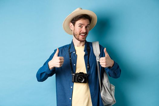 Happy smiling tourist say yes and show thumbs up, going on summer vacation, holding backpack and photo camera, blue background.