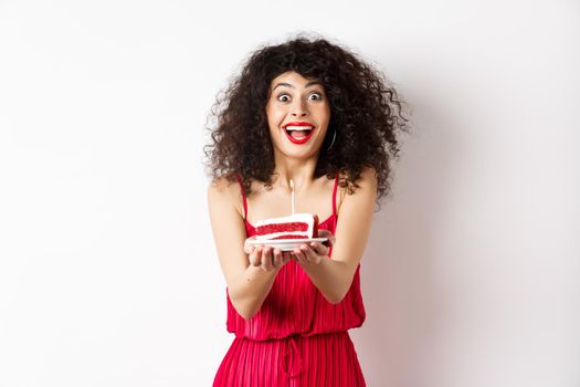 Holidays and celebration concept. Smiling woman in red dress giving you birthday cake with candle, standing over white background.