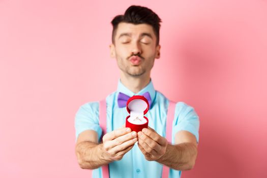 Valentines day. Silly boyfriend in bow-tie pucker lips, close eyes and waiting for kiss after making proposal, showing golden engagement ring in small red box, standing on pink background.