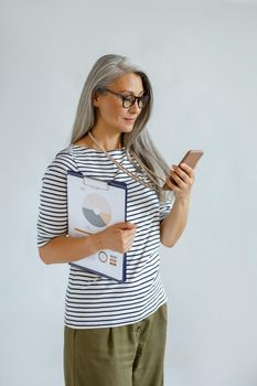 Positive mature Asian businesswoman in casual clothes holds diagrams and mobile phone standing on light grey background in studio