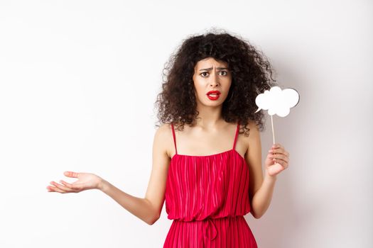 Confused young woman in red dress, frowning and looking upset, holding comment cloud stick, standing against white background.