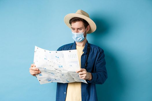 Covid-19, pandemic and travel concept. Tourist looking at map with sightseeing on vacation, wearing summer hat and medical mask from coronavirus, blue background.