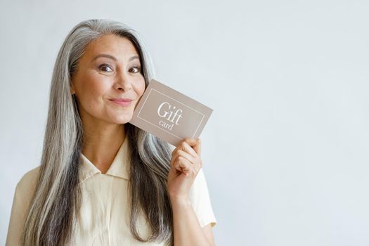 Joyful grey haired Asian woman holds gift card near face posing on light grey background in studio, space for text. Shopping certificate