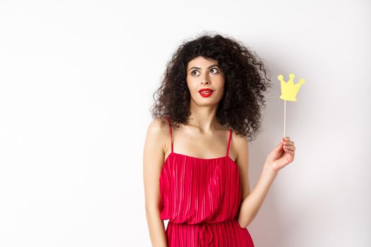 Stylish woman with curly hair in red dress, holding queen crown on stick and looking aside, standing on white background.