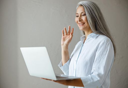 Smiling hoary haired mature Asian businesswoman waves hand to colleagues at videocall via modern laptop posing near grey stone wall in studio