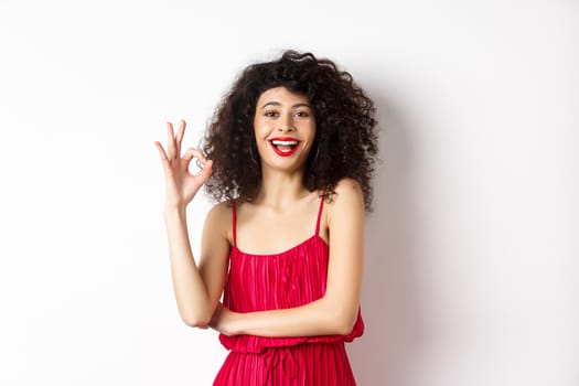 Cheerful caucasian woman with curly hair and makeup, wearing elegant red dress, showing OK sign and smiling in approval, standing over white background.