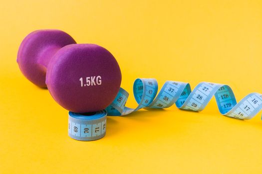 1.5 kilogram purple dumbbell and blue measuring tape on a yellow background, the dumbbell is based on a rolled up tape, copy space, weight loss concept