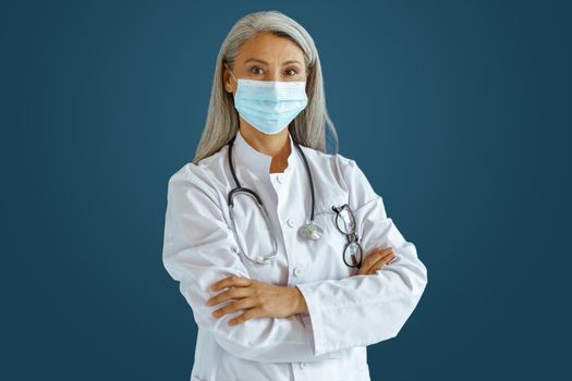 Grey haired Asian female doctor in white robe with medical mask and stethoscope stands on blue background in studio. Professional medical staff