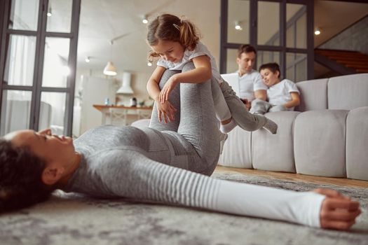 Low angle of smiling mother swinging girl on legs while boy is using notebook with dad on sofa