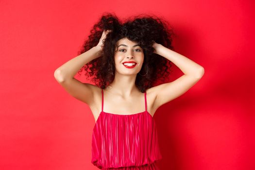 Carefree lady in red dress, touching her curly hair and smiling happy, standing on studio background. Copy space