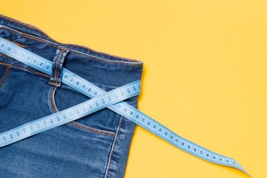 blue measuring tape is threaded into jeans instead of a belt, blue jeans on a yellow background, copy space, weight loss and weight control concept, measuring body volume