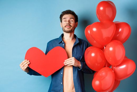 Valentines day concept. Man dreaming of true love, holding red heart cutout and standing near romantic balloons, blue background.