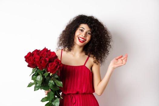 Happy woman celebrating, wearing stylish dress and holding flowers, smiling at camera, white background.