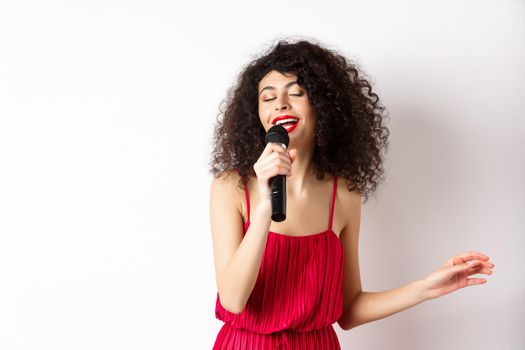 Happy elegant woman in red dress performing with microphone, singing karaoke and smiling, standing over white background.