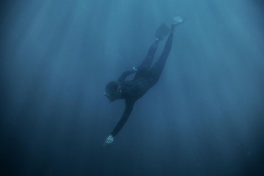 Sporty young man freediver wearing in wetsuit and flippers diving underwater in blue deep sea.