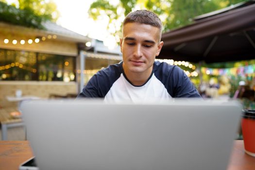 Young man sitting at table and typing on laptop keyboard while working in outdoor cafe, close up