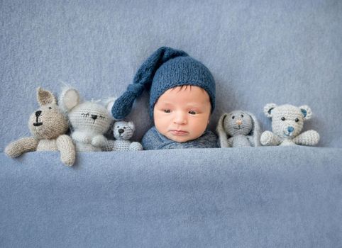 Cute newborn boy lying under blue blanket with plush toys near him, top view