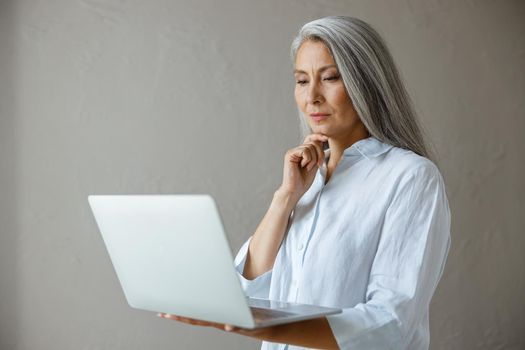 Attentive grey haired mature Asian lady in elegant blouse looks at modern laptop screen posing near stone wall in studio