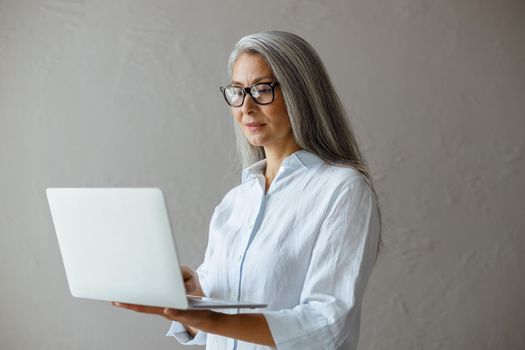 Concentrated long haired mature Asian woman wearing white blouse works on contemporary laptop standing near grey stone wall in studio