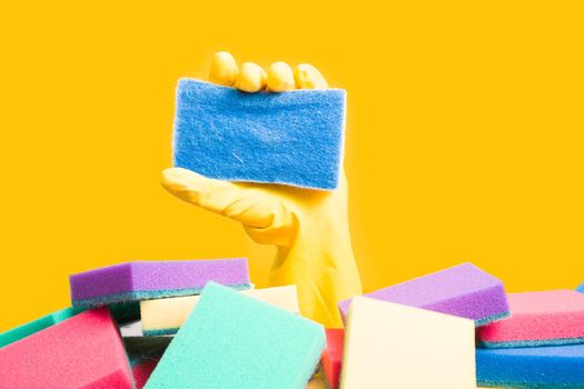 a hand in a yellow rubber glove holds a sponge for washing dishes and cleaning, a hand sticks out of a pile of sponges, yellow background copy space