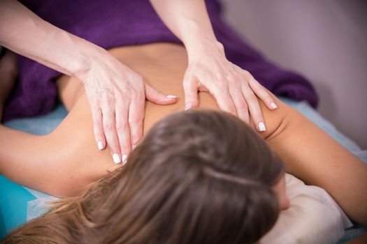 Close-up of back of the young skinny girl with tanned skin with closed eyes lying in bed and relaxing while getting back massage at the clinic, close-up of the masseur's hands and girl's back