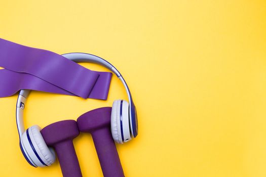 Sports equipment,gray and blue headphones and purple dumbbells on a yellow background, purple rubber expanders, copy space, top view, sport and music concept