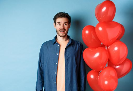 Happy valentines day. Excited smiling guy standing near red hearts balloons and looking at camera, blue background.