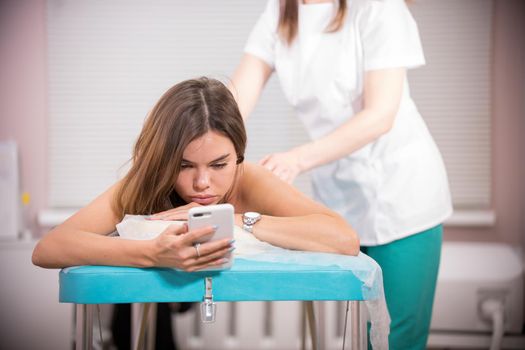 Masseur doing a back massage to a young skinny woman with tanned skin looking at her phone screen at the clinic, close-up of the patient's face