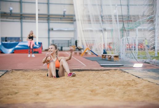 Young blonde smiling women performing a long jump in the sports arena. Sitting in the sand. Mid shot