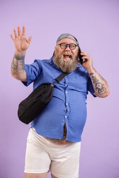 Jouful mature plump man with crossbody bag talks by mobile phone and waves hand standing on purple background in studio