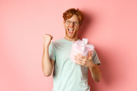 Valentines day and romance concept. Cheerful redhead guy making fist pump and scream yes with joy, holding gift box, smiling, standing over pink background.